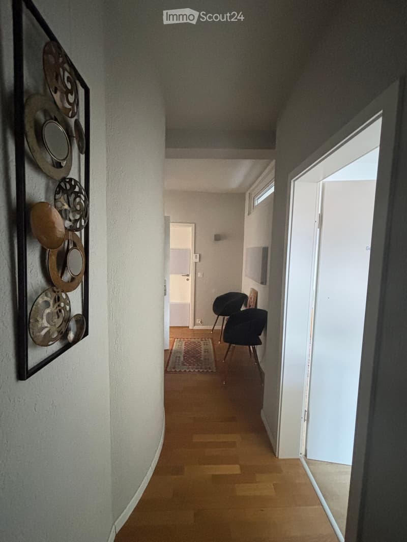 A curved hallway with a wall decoration, wooden flooring, and an open doorway leading to a door.