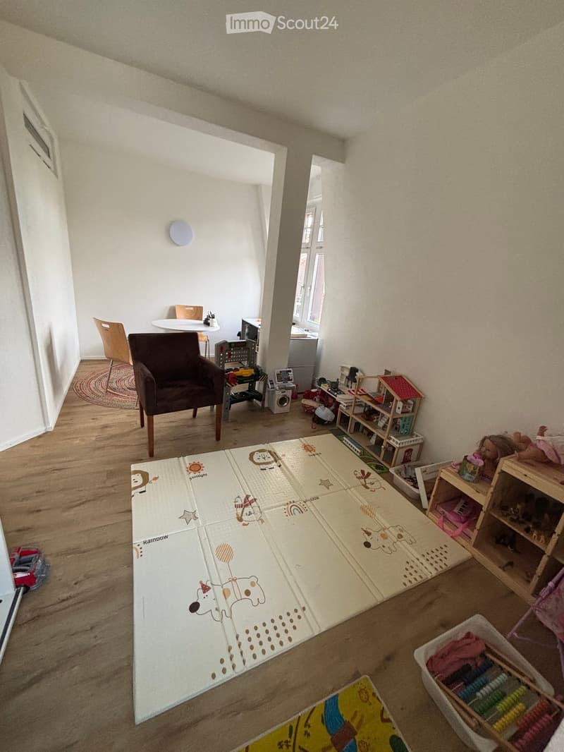 A playroom with a large playmat, a wooden chair, a small table, a white desk, shelves with toys, and a rug