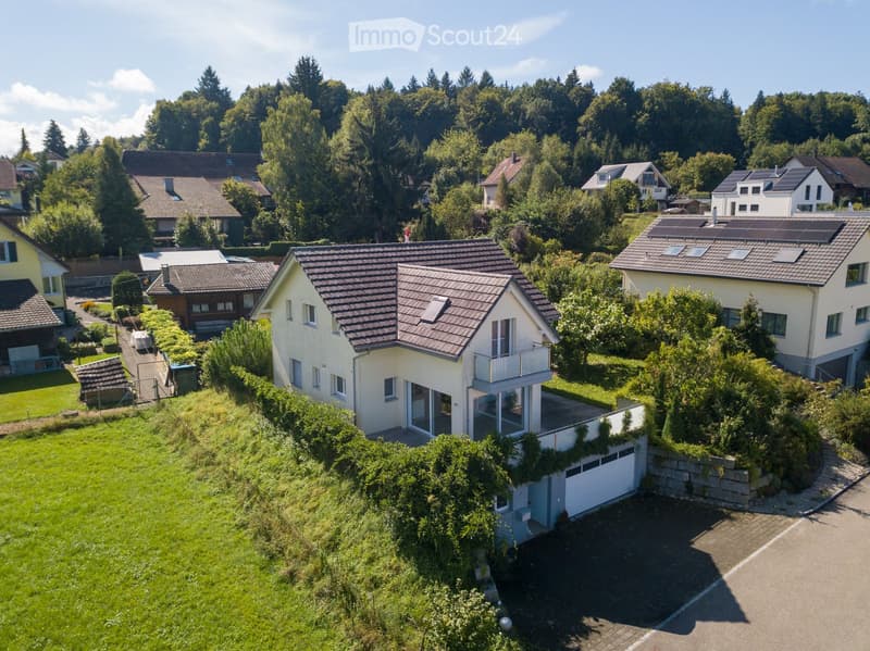 Detached house, brown roof, solar panels, white garage, situated on the slope of a hill, surrounded by greenery