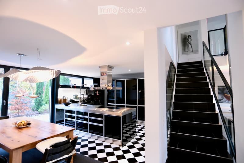 a modern interior of kitchen with black and white checkered flooring, central island with stove, a modern staircase, open glass door, dining table with chairs