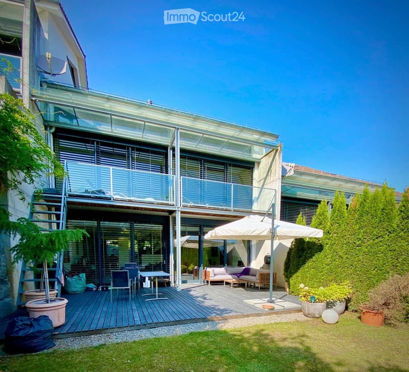 Modern architecture, large windows, balcony with glass railings, outdoor seating area, lush green garden, outdoor dining table, hedges, trees