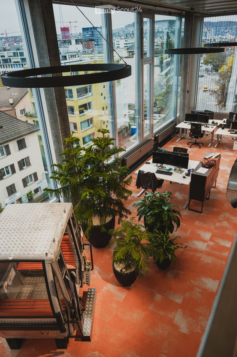 Open floor plan, large windows, desk with monitors, chairs, and potted plants, office vehicle parked