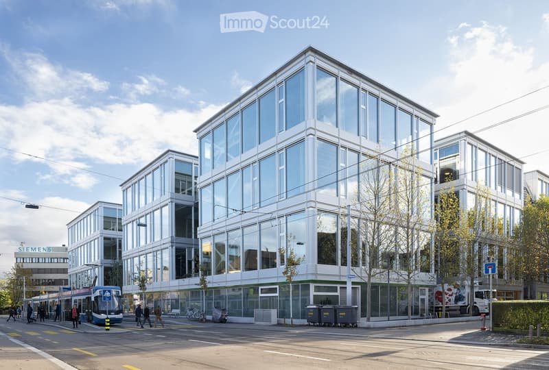 Modern office building with glass walls, large glass windows, multiple floors, city street with a tram, trash bins, and parked bikes.