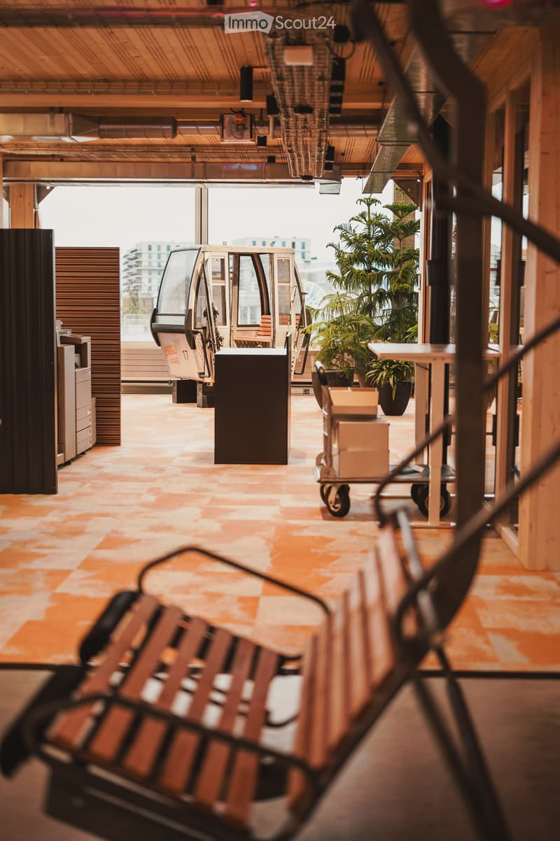 a room with a tile floor, wooden lounge chair, table, plants, office machines, a lift car, open windows, and a clear sky