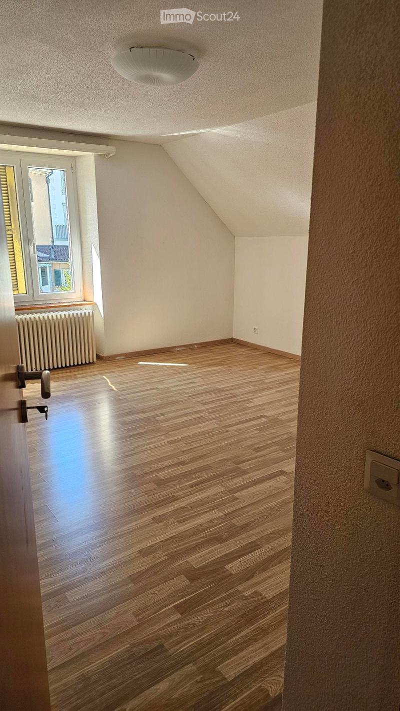 A empty room with wooden flooring, white walls, a radiator, a window with yellow shutters, and a door with a lock.