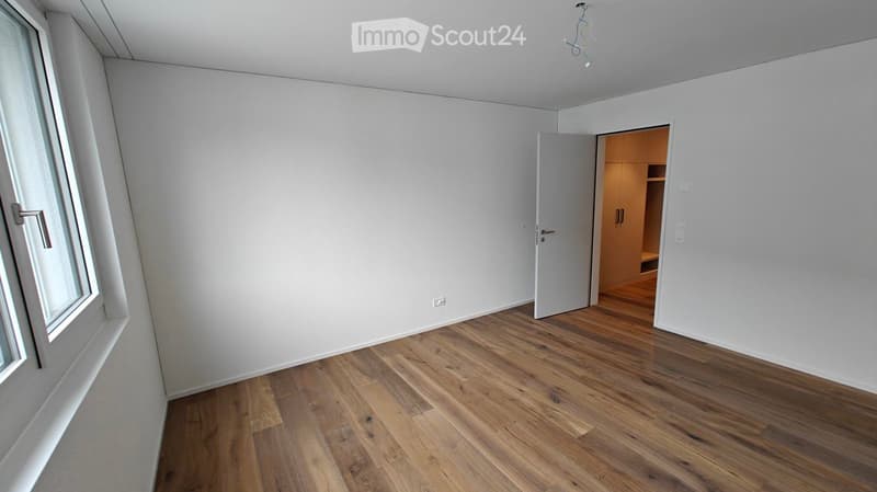 Empty room with light wooden flooring, white walls, and a large window. An open closet with wooden shelves is visible in the background.
