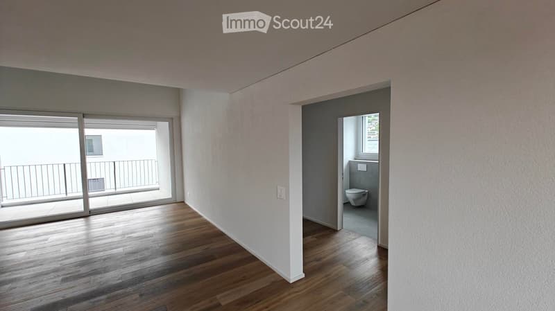 Empty room with wooden flooring, white walls, and a sliding glass door leading to a balcony. A small bathroom is visible through an open doorway.