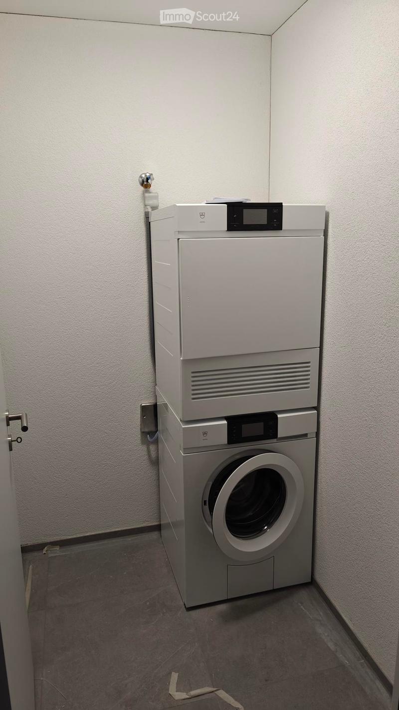 A small room with a white wall and a gray floor. There is a white washing machine and a white dryer stacked on top of each other in the corner of the room. The washing machine has a digital display on top and a door handle on the front. The dryer has a di