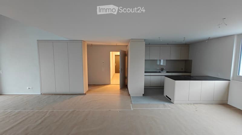 Empty room with white walls, wooden floor, kitchen area with island, cabinets, and sink, large window, and a door leading to another room.