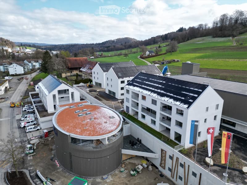 Aerial view of a modern building complex with a circular structure, surrounded by a green field and mountains in the background.