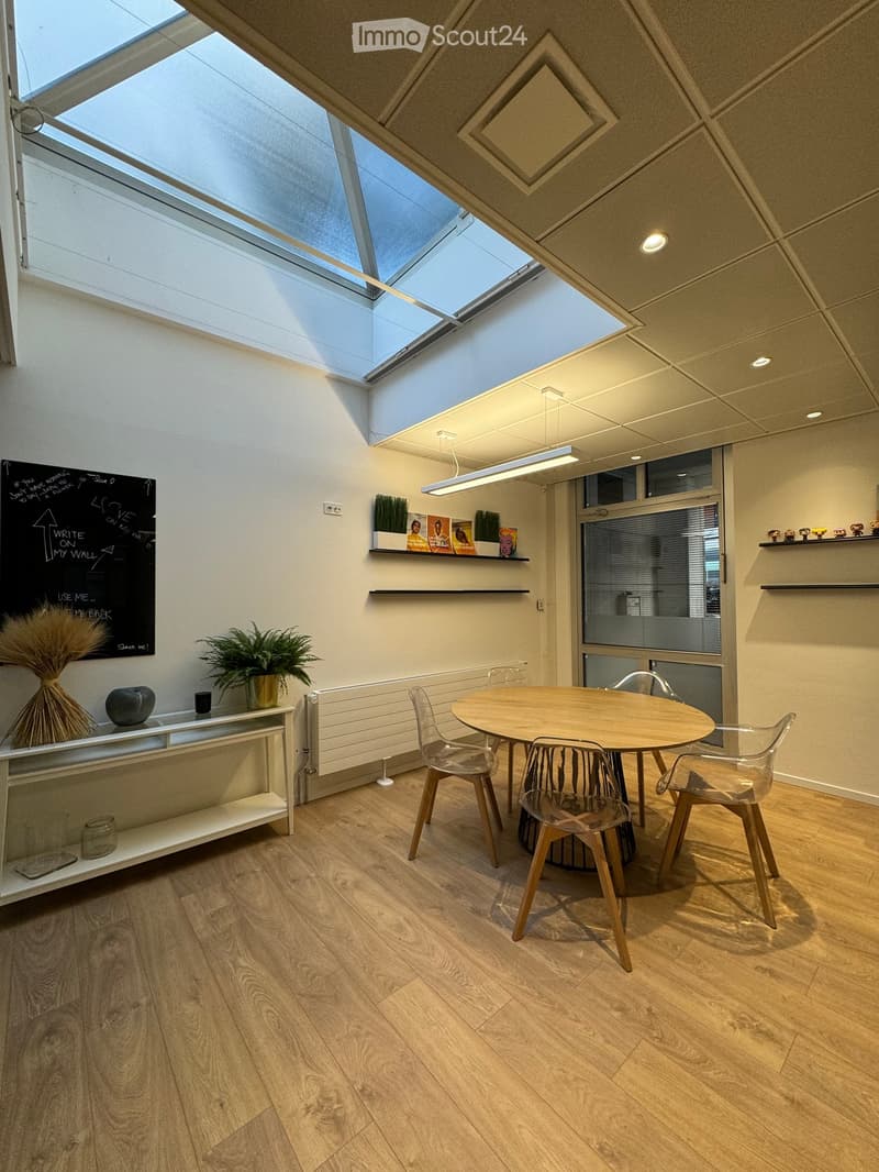 The room features a round wooden dining table with four plastic chairs. There is a glass door with a blind on the side. On the opposite wall, there is a chalkboard and a white shelf with a plant and decorative items. Above the table, there is a hanging la