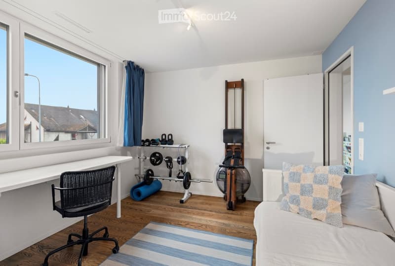 A room with a desk, chair, weight set, and yoga mat. The room has hardwood floors, a window with a blue curtain, and a white bed with pillows. The walls are painted blue and white, and there is a rug on the floor.