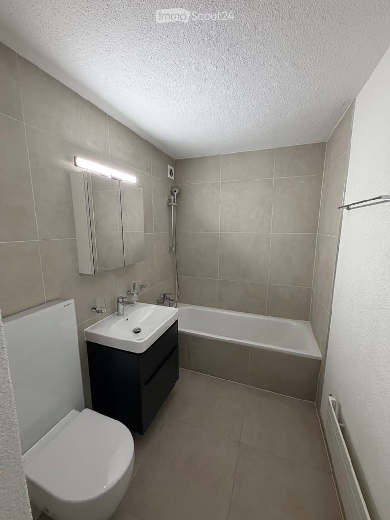 Modern bathroom with gray tiled walls, white bathtub, white sink with mirror, toilet, and overhead lighting