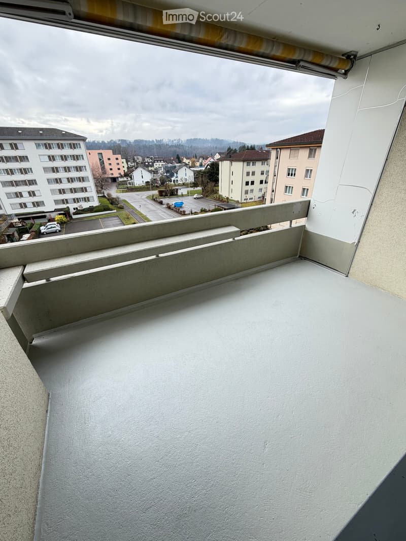 Balcony with a grey floor, a grey wall, and a metal railing. View of the parking lot and several buildings.