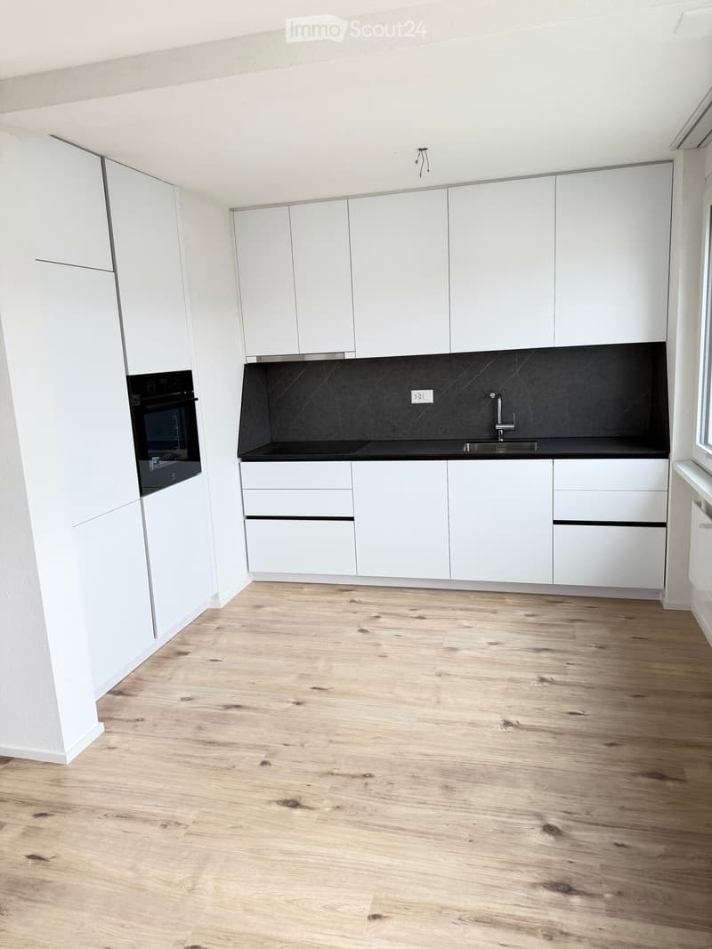 Empty room with white kitchen cabinets, black counter, white walls, wooden floors, integrated dishwasher, washing machine, built-in oven, sink and faucet.