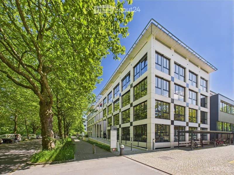 large multistory office building with glass facade, located in a well-kept urban area, surrounded by greenery and trees, with a paved sidewalk, a car park, and a bicycle stand