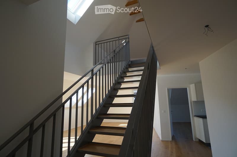 Interior view of a modern building featuring a sleek staircase with dark grey handrails, wooden steps, and an open, airy layout illuminated by natural light.