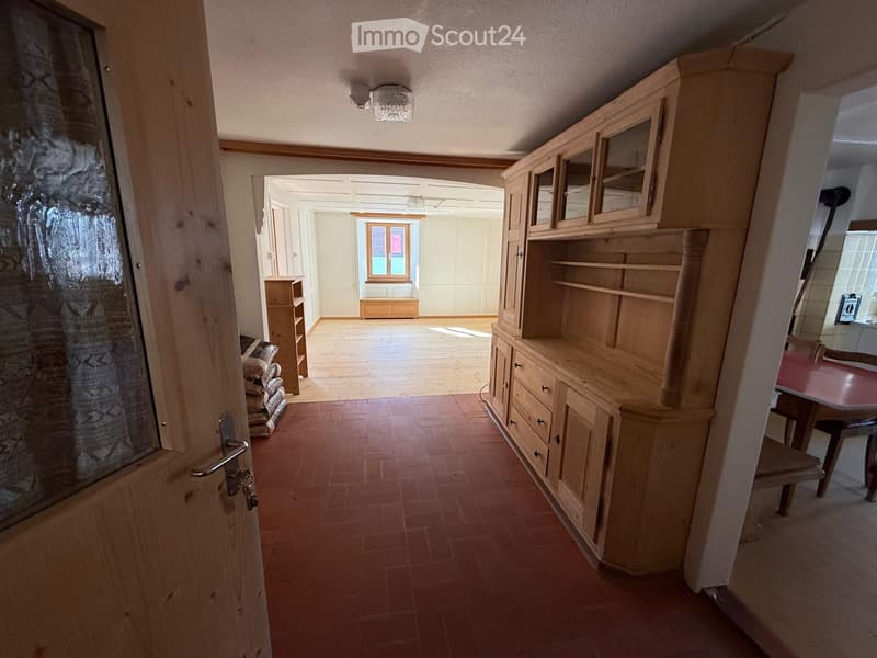 a wooden door leads into a hallway, a large wooden shelf is in the hallway, an arched door leads to a room with a window, a wooden table and chairs are to the right, the hallway has a tiled floor
