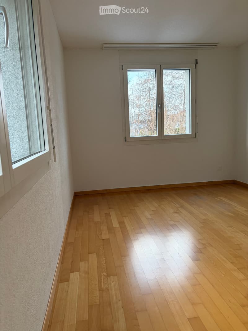 Empty room with wooden flooring, light-colored walls, and large windows allowing natural light to enter.