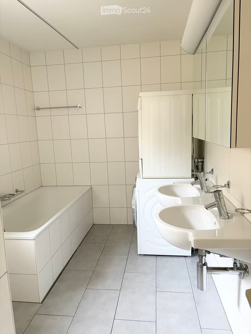 Modern bathroom with white tiled walls and floor, featuring a bathtub, double sink vanity with chrome faucets, and a large mirror above the sinks.