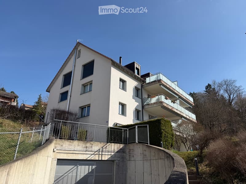 A modern two-story house with white walls, multiple windows, and a balcony on the upper level. The house is situated on a raised ground floor with a concrete staircase leading up to it.