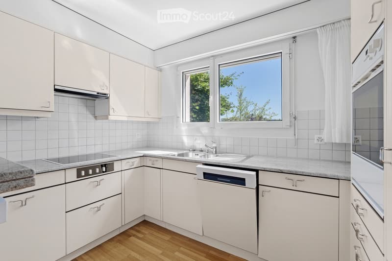 Modern kitchen with white cabinets, granite countertops, a sink, a dishwasher, and a stove. The room features wooden flooring and large windows with white curtains.