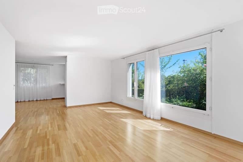 Bright, empty room with light wooden flooring and white walls. Large windows with white curtains allow natural light to fill the space. The room features a sliding glass door leading to an outdoor area with greenery visible.