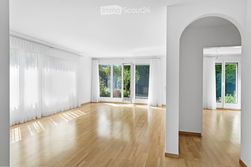 Bright, unfurnished room with light wooden flooring, large windows with white curtains, and an arched doorway leading to another space.
