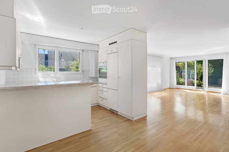 Bright, modern kitchen with white cabinets, wooden flooring, and large windows overlooking a garden. Features a central island, built-in oven, and ample natural light.
