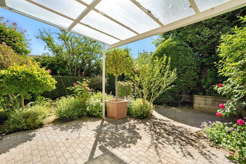 A well-maintained garden area with a paved brick floor, lush greenery, flowering plants, and a potted tree under a pergola with a translucent roof.
