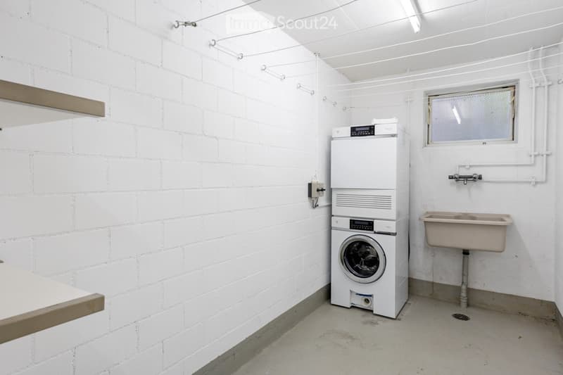 A small, white-walled utility room with a washing machine and a sink, featuring exposed brick walls and a window with a metal mesh cover.