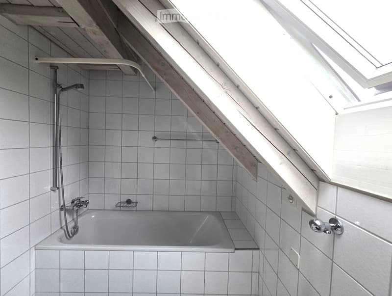 Bathroom: white tiled floor and walls, tub, ceiling slanted due to attic, mounted shower head, metal handrails, ventilation window