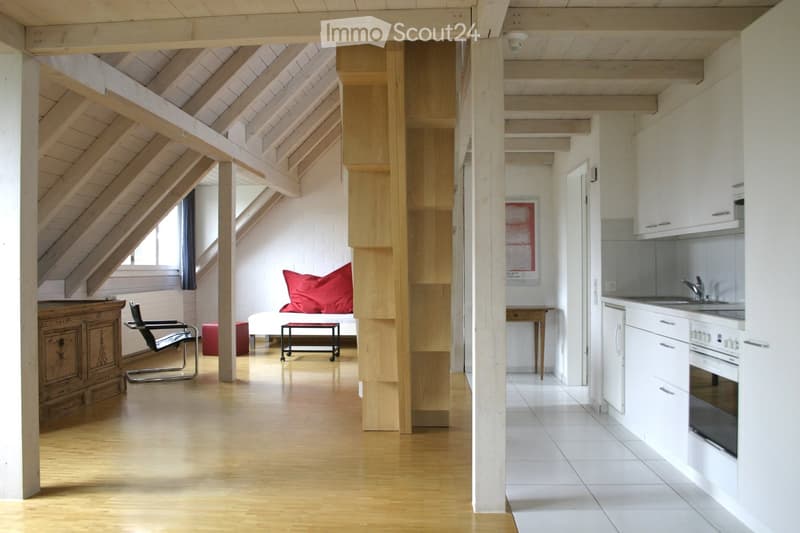 A wooden staircase, a white kitchen with white cabinets and tiles, a wooden table with a red pillow, a wooden chair, a bed, and a window.