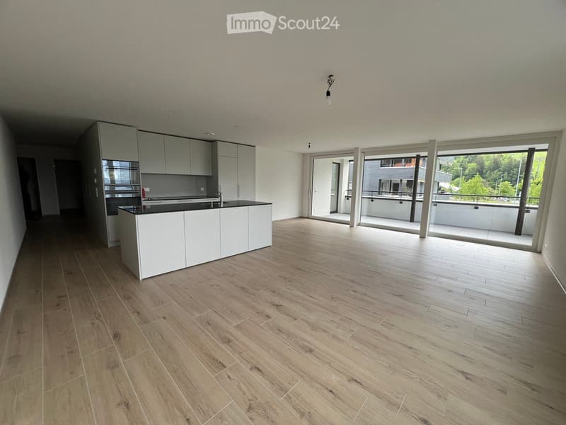 Empty kitchen with white cabinets, black countertop, and wooden flooring, leading to a balcony area with a view of greenery.