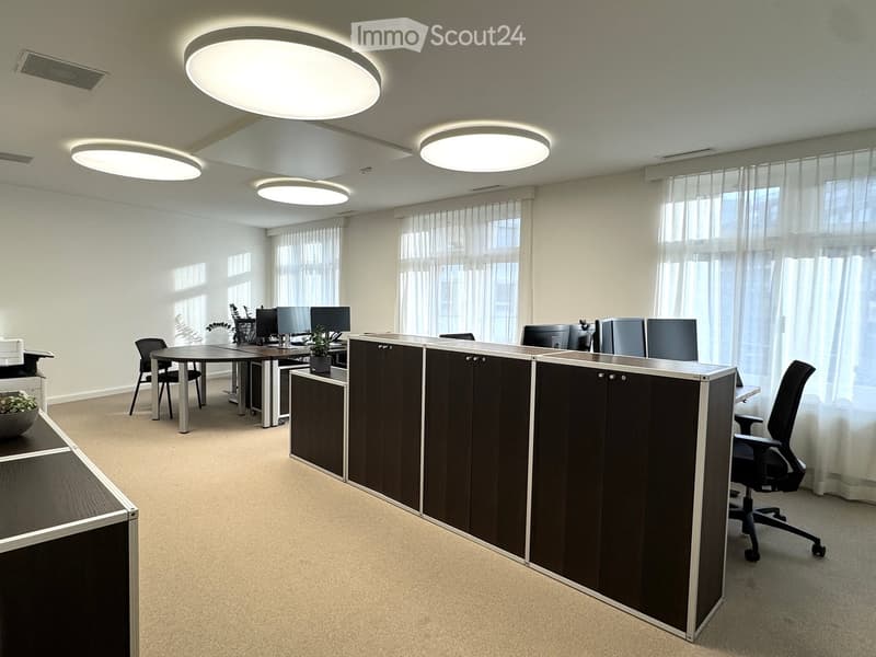 Modern office interior with beige carpet, beige walls, and white ceiling, equipped with a large desk, computer setup, office chairs, round table, a black cabinet, and multiple round ceiling lights.