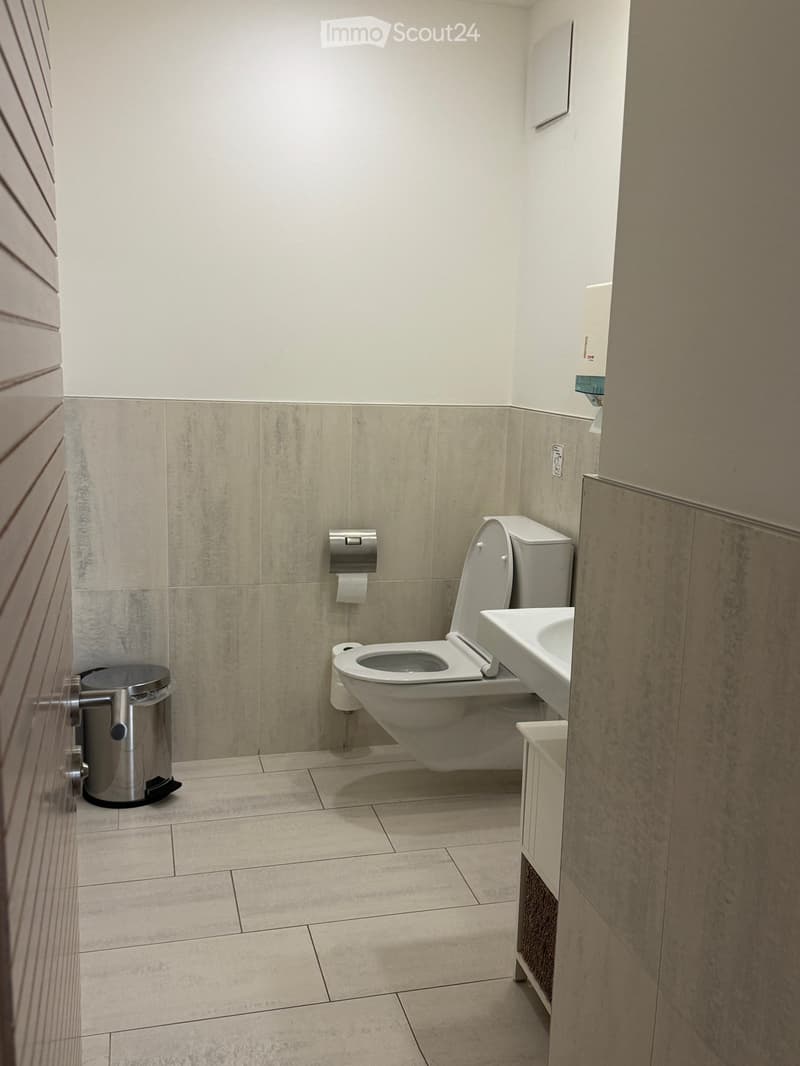 Compact bathroom with white tile floor, wall-mounted toilet, vanity with sink, and a silver trash bin.