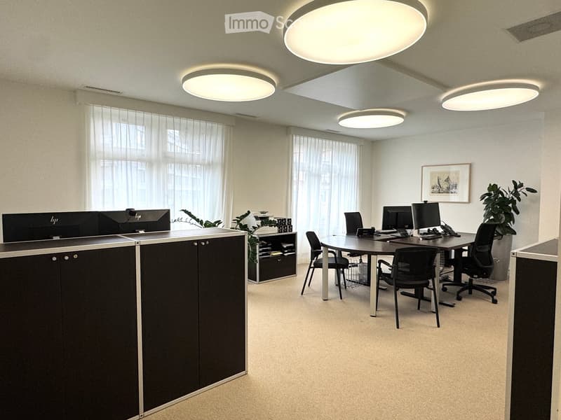 Modern office space with contemporary furniture, LED ceiling lights, white walls, and large windows with curtains. The room features a round desk with chairs, a printer, and a potted plant.