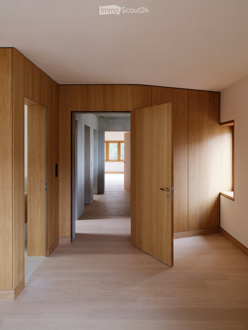 Modern hallway with light wooden doors and walls, light wooden flooring, and a view into another room with a large window.