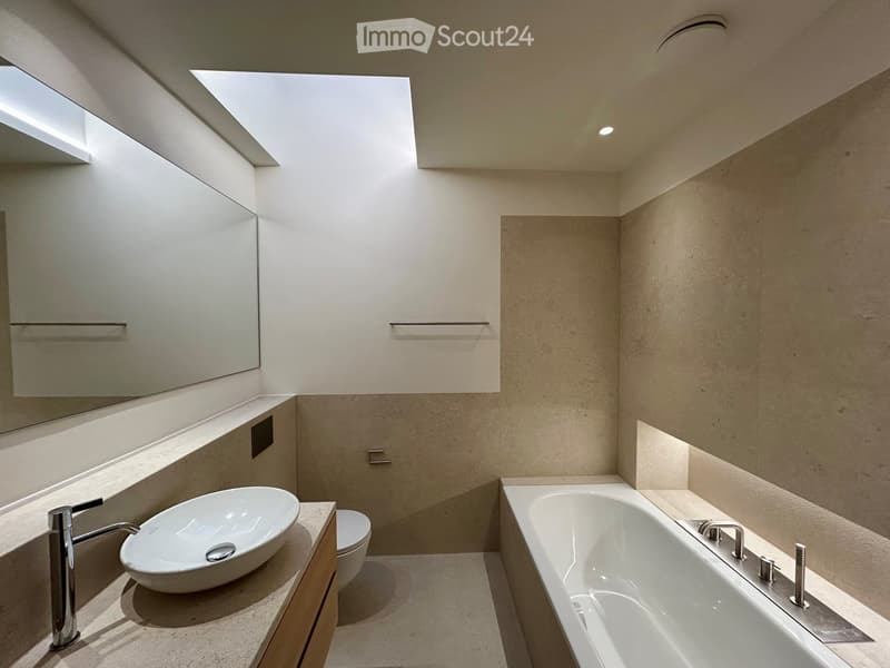 Contemporary bathroom with skylights, a large mirror, wooden vanity, white sink, toilet, bathtub with shower faucets, built-in cabinet, beige tiled walls, and ceiling lights.