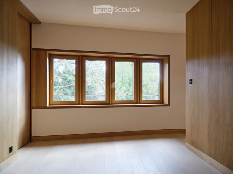 Empty room with light wooden flooring, beige walls, and large windows with wooden frames, offering a view of greenery outside.