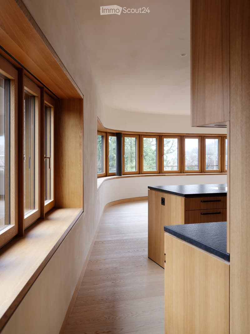 Modern kitchen with light wood cabinetry, sleek black countertops, and large windows offering a view of greenery.