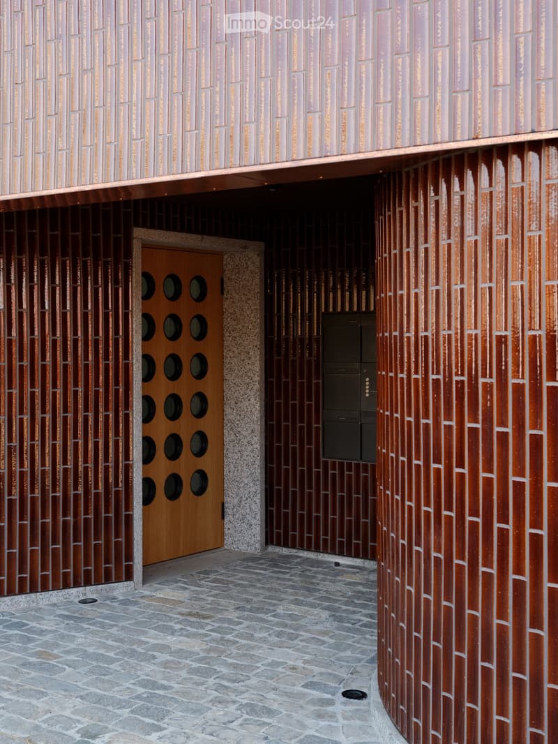 Entrance area of a modern building with a textured brick facade, a wooden door with circular cutouts, and a stone-paved ground.