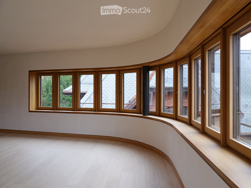 Empty room with light wooden flooring, curved walls, and large wooden-framed windows offering views of the roof and surrounding greenery.