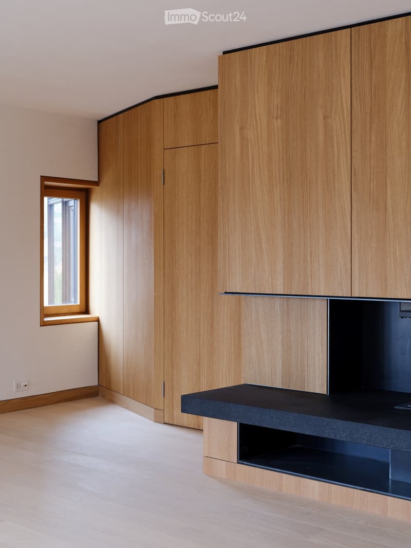 Empty room with light wooden flooring, light-colored walls, and built-in wooden cabinetry with a black countertop. A small window is visible on the left side.