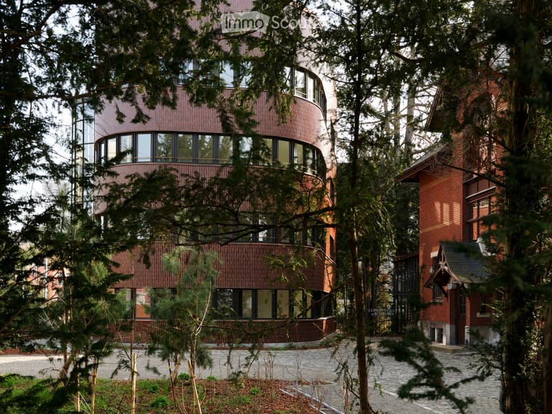 A modern, two-story house with a curved facade, large windows, and a brick exterior, surrounded by trees and a stone pathway.
