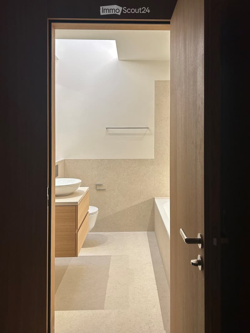 Modern bathroom with a sink, toilet, bathtub, and wooden cabinet. Light-colored tiles and a sleek design.