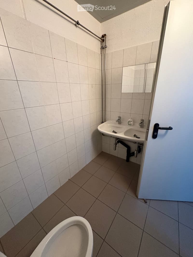 A small bathroom with white tiled walls, tiled floor, a sink with two faucets, a mirror, and a toilet.