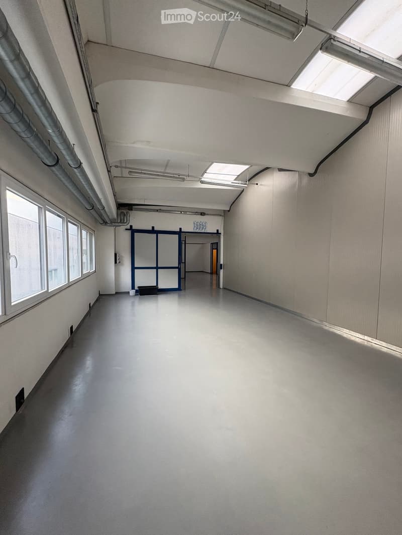 Empty room with white walls, gray floor, large windows, metal pipes on ceiling, double doors, and a small door at the end.