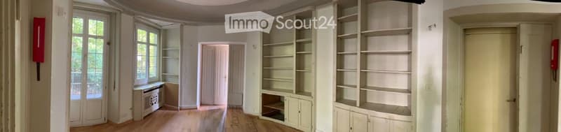 Empty room with wooden floor, white walls, large windows, built-in shelves, and a door.
