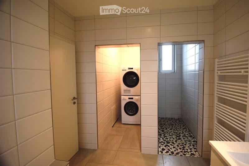 A small room with a washing machine and a dryer, a small window, and a shower area with a towel rack.